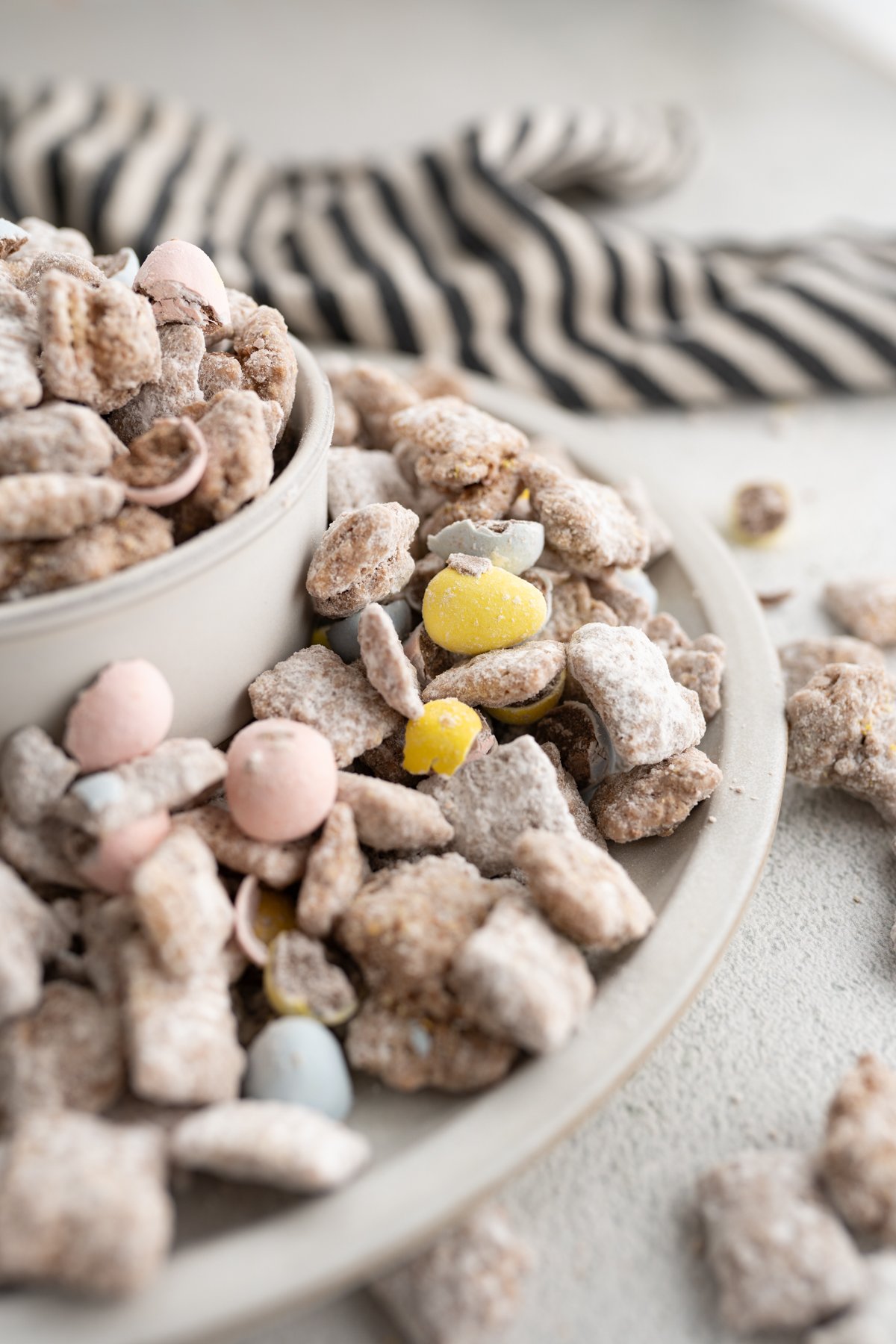 Side view of a bowl of cadbury egg muddy buddies overflowing onto a plate. 