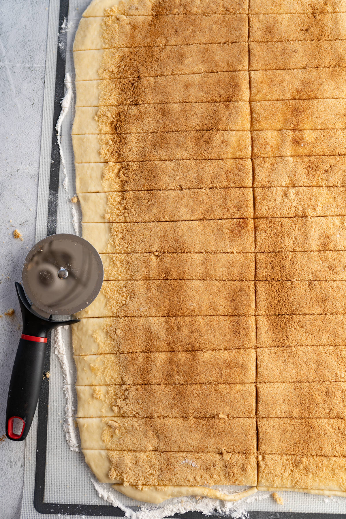A pizza cutter sitting next to a row of cinnamon roll dough before being rolled up.