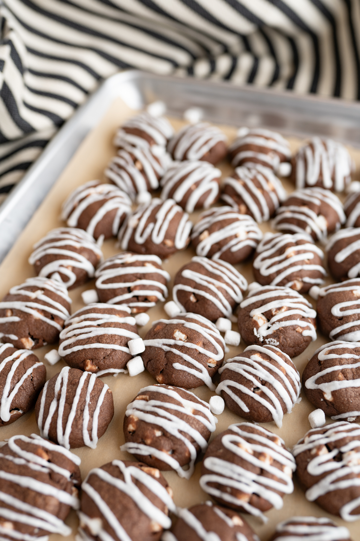 Hot chocolate cookies, stuffed with mini marshmallows and drizzled with white chocolate on a cookie sheet. 